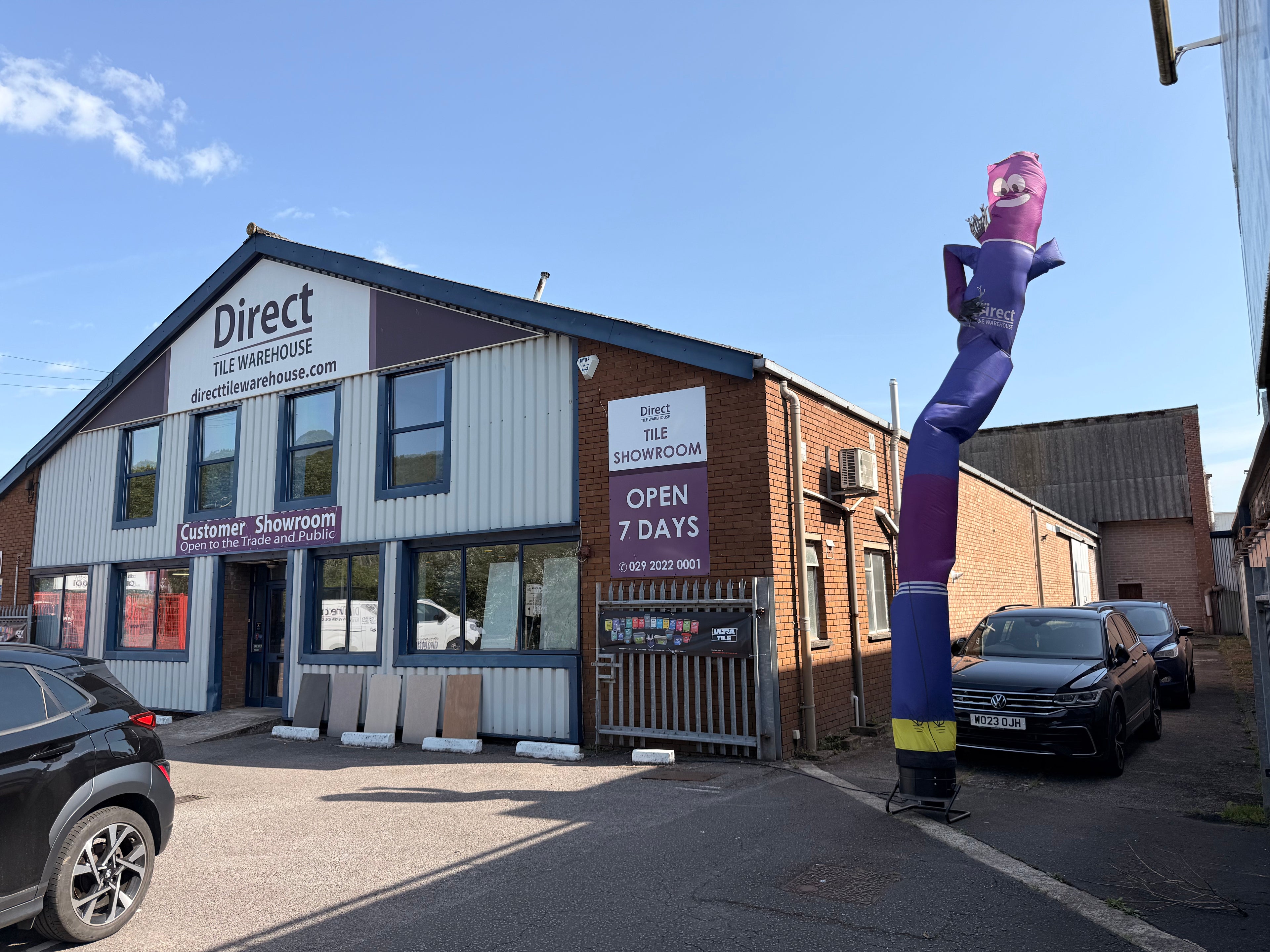 Direct Tile Warehouse showroom exterior under a blue sky, featuring a brick and corrugated metal building with a large purple inflatable air-traffic controller on the sidewalk and parked cars.