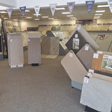 A showroom interior with multiple tile and flooring sample displays, price tags, hanging pennant banners, and a carpeted floor. No person is visible.